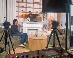 Two people seated on green sofas recording a podcast or interview in a studio, with microphones, cameras on tripods, studio lights, and a decorative fireplace in the background.