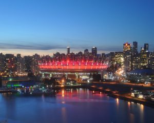 Vancouver skyline at dusk with BC Place stadium illuminated in red, reflected in the water, and surrounded by city buildings.