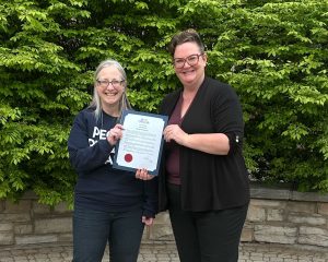 Two people standing outdoors smiling and holding a framed certificate, with green bushes in the background.