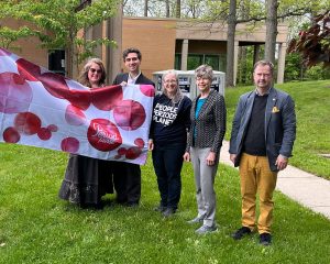 Five people standing outside on grass holding “The Period Purse” flag, with a building and trees behind them.