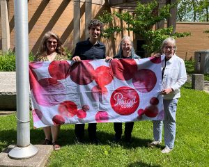 Four people standing outdoors on a lawn holding a large “The Period Purse” flag with red and pink circular patterns.