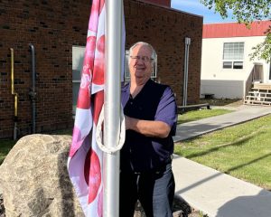 An older person outdoors raising “The Period Purse” flag on a flagpole beside a brick building on a sunny day.
